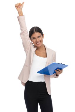 Pembe ceketli çekici esmer kadın elini havada tutuyor, başparmak hareketi yapıyor, panoyu tutuyor ve stüdyoda beyaz arka planda gülümsüyor.