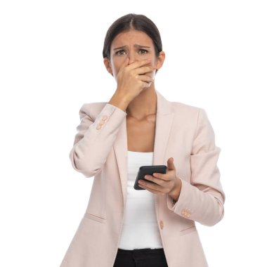 Pembe ceketli üzgün genç iş kadını mutsuz bir yüz ifadesi takınıyor, elleriyle ağzını kapatıyor ve stüdyoda beyaz arkaplan üzerine yazılar okuyor.