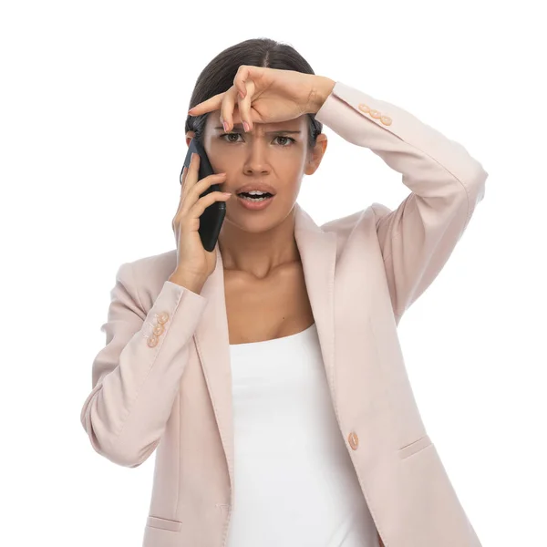 ピンクのジャケットを着た頭のいいカジュアルな若い女性が電話で話し額に手をつないでスタジオで白い背景に頭痛を抱えています — ストック写真