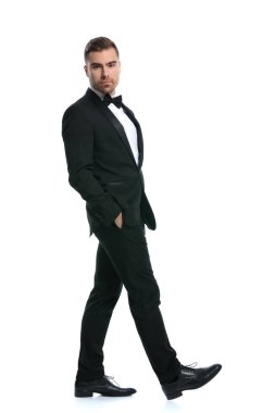 Siyah smokinli yakışıklı sakallı iş adamı ceplerinde el ele tutuşuyor ve stüdyoda beyaz arka planda tek başına yürüyor.