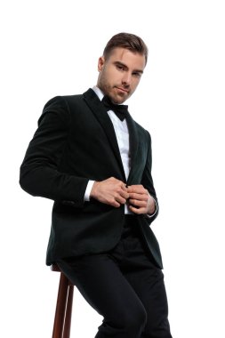 Ahşap sandalyede oturan, siyah smokin giyen ve stüdyoda beyaz arka planda poz veren mutlu genç bir adam.