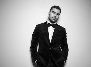 Siyah smokinli, papyonlu, el ele tutuşmuş stüdyoda kendine güvenen bir şekilde poz veren havalı bir genç. Siyah beyaz bir portre.