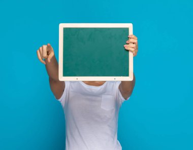 Sıradan bir kadın yeşil bir panonun arkasına saklanıyor ve mavi arka plana başparmak işareti yapıyor.