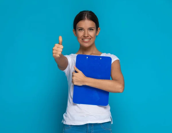 stock image beautiful casual woman giving a thumbs up and holding a clipboard against blue background