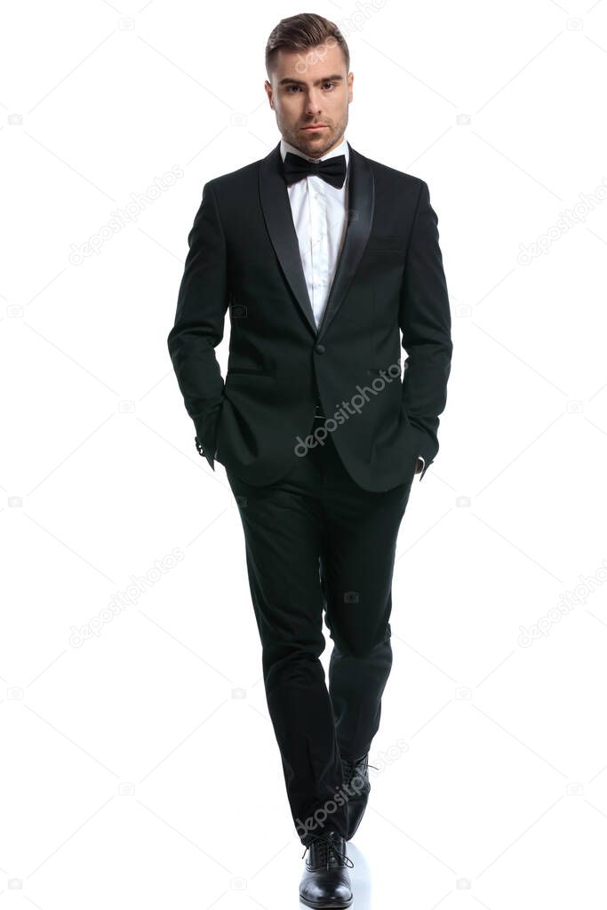 cool young businessman in black tuxedo holding hands in pockets and confidently walking isolated on white background in studio, full body picture