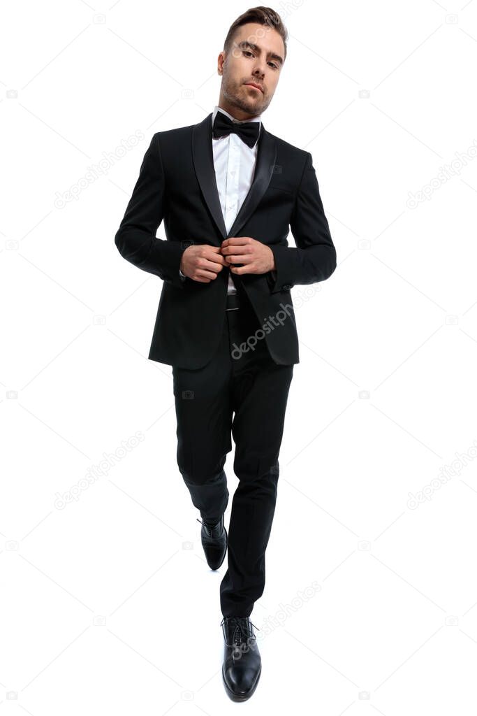 elegant young fashion man buttoning black tuxedo and walking isolated on white background in studio, full body picture