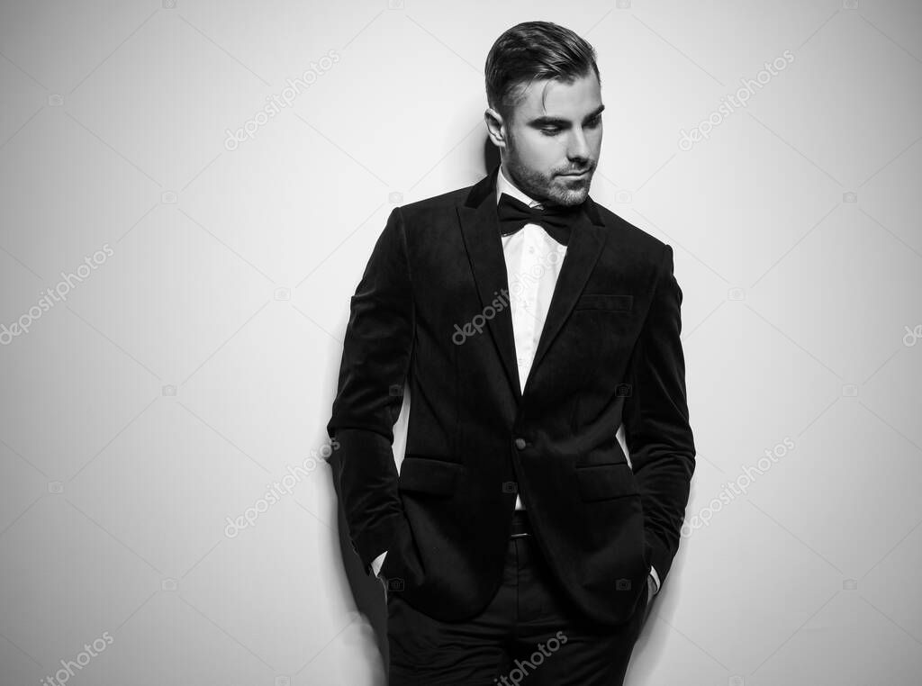 portrait of sexy young businessman in black tuxedo posing with hands in pockets and looking to side while standing in studio