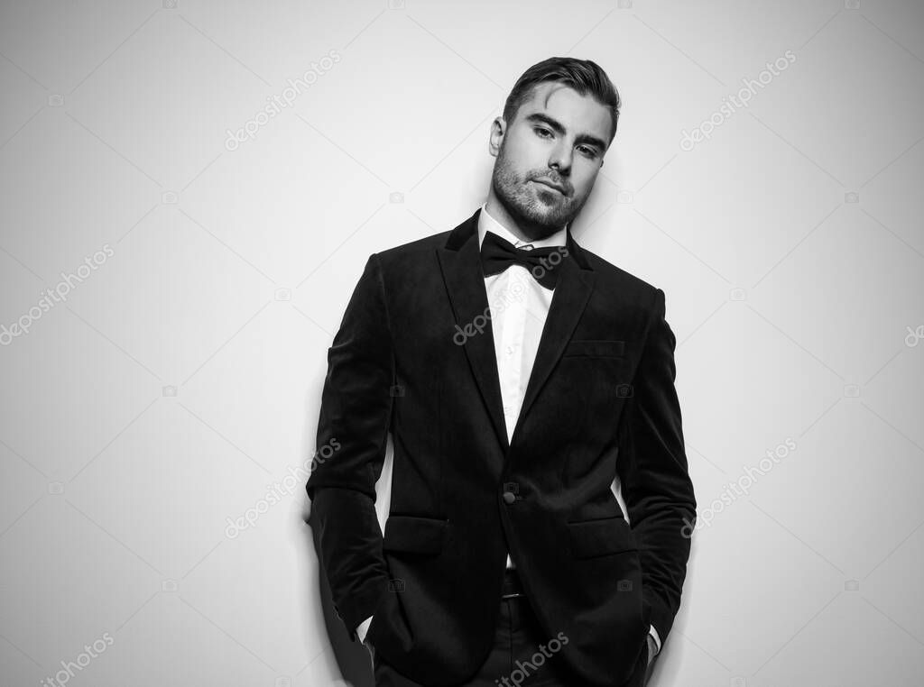 dramatic cool young man in black tuxedo with bowtie holding hands in pocket and posing in a confident manner in studio, black and white portrait