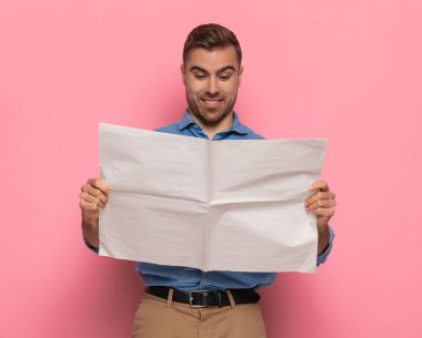 Heyecanlı sakallı adam gazete okuyor ve stüdyoda pembe arka planda poz verirken mutlu bir yüz ifadesi takınıyor.