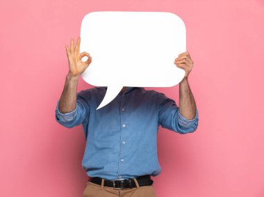 Kot gömlekli genç adam konuşma balonunun arkasına saklanıyor ve stüdyoda pembe arka planda iyi bir jest yapıyor.