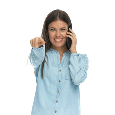 Genç, sıradan bir kadın kamerayı işaret ediyor ve telefonda beyaz arka planda konuşuyor.