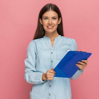 Elinde mavi bir pano tutan ve pembe arka planda kameraya gülümseyen sıradan bir kadın.