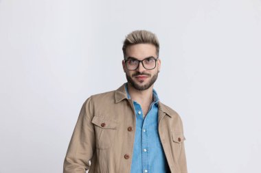 Gözlüklü, kot gömlekli, bej ceketli, gri arka plana bakıp gülümseyen mutlu, tıraşsız adam.