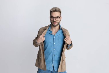 Kot pantolon giyen, gözlüklü, bej ceketli yakışıklı sakallı adam. Stüdyoda gri arka planda seksi duruyor ve poz veriyor.