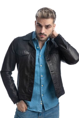 Deri ceketli havalı adam el ele tutuşuyor. Boynunu ve cebini tutuyor. Stüdyoda beyaz arka plana karşı güvenle poz veriyor.