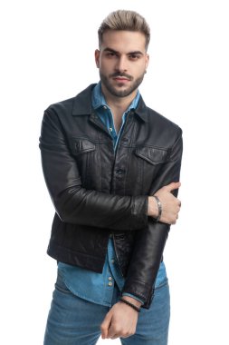 Deri ceketli, kolları moda pozunda gezen, gülümseyen ve stüdyoda beyaz arka planda poz veren genç bisikletçi.