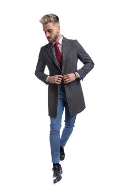 Havalı, takım elbiseli genç bir işadamı uzun ceketini ilikleyip stüdyoda beyaz arka planda tek başına yürürken tüm vücudunu gösteriyor.