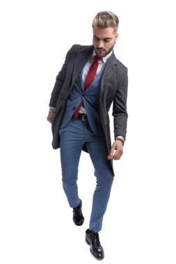 Mavi takım elbiseli, uzun paltolu, beyaz arka planda yürürken kendine güvenen şık bir moda adamının tüm vücudu.