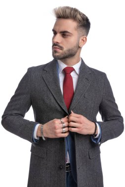 Mavi takım elbiseli traşsız genç adam uzun paltosunu kapatıyor ve beyaz arka planda tek başına dikilirken yan tarafa bakıyor.