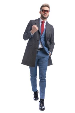 Takım elbiseli, uzun ceketli, güneş gözlüklü, el ele tutuşmuş bir iş adamı. Stüdyoda beyaz arka planda tek başına yürürken kutlama yapıyor.