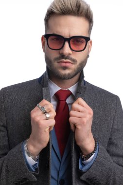 Takım elbiseli, güneş gözlüklü, uzun yakalı ve stüdyoda beyaz arka planda poz veren şık bir adamın portresi.