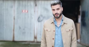 Tıraşsız, seksi bir adam, bej ceketini çekiştirirken ve düzeltirken kendine güvenen bir şekilde yürüyor. Dışarıdaki gri metal garajına bakıp gülümsüyor.