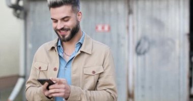 Kot gömlekli mutlu yakışıklı adam e-posta okuyor, telefonlara cevap veriyor, baş parmak hareketi yapıyor ve gri metal garajın önünde gülümsüyor.