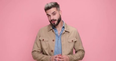 Kendine güvenen havalı adam, bej ceketli, parmaklarını ovuşturup gülümsüyor, silah yapıyor ve stüdyoda pembe arka planda ateş ediyor.