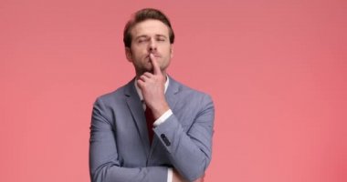Zarif takım elbiseli düşünceli, mutlu bir adam etrafa bakıyor, düşünüyor, bir fikri var, gülümsüyor ve stüdyoda pembe arka planda güzel bir jest yapıyor.