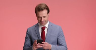 Kırmızı kravatlı mutlu iş adamı telefon tutuyor ve mesajlar yazıyor, stüdyoda pembe arka planda başını sallıyor ve gülümsüyor.