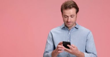 Şaşırmış genç bir adam telefona sarılıyor, iyi bir tartışma yapıyor, stüdyoda pembe arka planda işaret ediyor ve gülüyor.