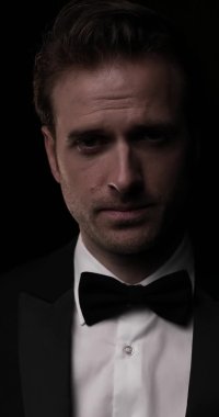 Genç ve seksi bir işadamının kameraya baktığı dikey bir video, aşağı bakıyor, smokini tamir ediyor ve gülümsüyor.