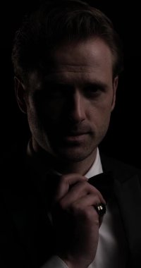 Papyonunu ve smokinini ayarlayan, karanlık arka planda kameraya gülümseyen genç bir iş adamının dikey videosu.