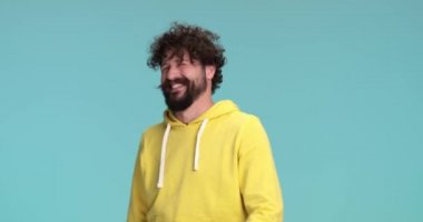 Kıvırcık saçlı ve uzun sakallı komik adam eğleniyor, şakalar yapıyor ve gülüyor, kollarını havaya kaldırıyor ve stüdyoda mavi arka planda neşeli hareketler yapıyor.