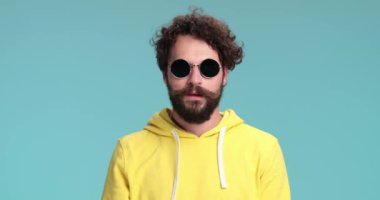 Uzun sakallı ve bıyıklı kıvırcık saçlı adam surat yapıyor, bıyık bırakıyor, eğleniyor, gülümsüyor ve stüdyoda mavi arka planda surat asıyor.