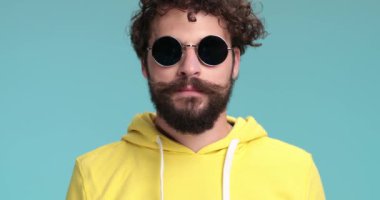 Kıvırcık saçlı, güneş gözlüklü, sakalını kaşıyan ve elleriyle gülümseyerek stüdyoda mavi arka planda olmaktan gurur duyan mutlu genç bir adam.