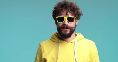 Havalı güneş gözlüklü mutlu sakallı adam, sarı kazak giyiyor, gülüyor, konuşuyor, başını sallıyor ve el hareketi yapıyor stüdyoda mavi arka planda dururken.