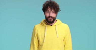 Uzun sakallı, sarı kapüşonlu üzgün bir genç adam üzgün bir yüz yapıyor, şans diliyor ve stüdyoda mavi arka planda poz veriyor.