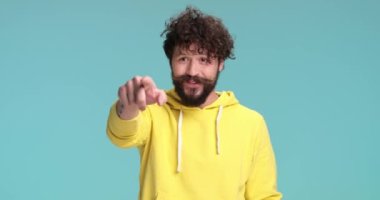 Sarı kapüşonlu mutlu adam el sallıyor, parmağıyla işaret ediyor ve davet ediyor, yumruklarını kaldırıp neşeleniyor, gülümsüyor ve mavi arka planda stüdyoda eğleniyor.