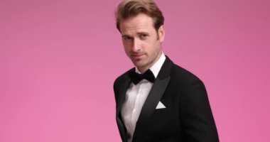 Siyah smokinli çekici, tıraşsız damat kendine güvenen, gülümseyen, yandan bakan ve stüdyoda pembe arka planda papyon takan.