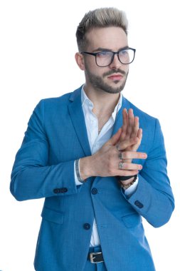 Tıraşsız, gözlüklü, kravatsız, mavi takım elbiseli genç bir işadamı, beyaz arka planda stüdyoda poz verirken avuçlarını bir yandan ovuyor, bir yandan da avuçlarını ovuyor.