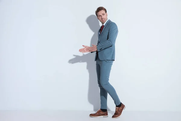 青いスーツを着たセクシーなビジネスマンが側を見て手のひらをこすりスタジオで灰色の背景を歩く姿を — ストック写真