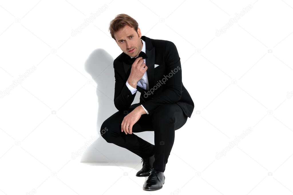 handsome businessman squatting, fixing his bowtie and looking at the camera on white background