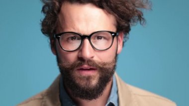 surprised young bearded guy making a shocked face, opening mouth and getting good news, smiling and positive nodding on blue background in studio