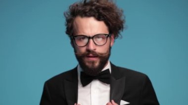 sexy long bearded guy with moustache wearing tuxedo and eyeglasses, making a snapping finger gesture and nodding against blue background in studio