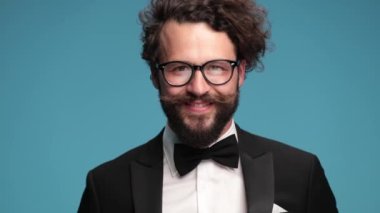 attractive elegant man in black tuxedo with bowtie smiling, fixing and adjusting tuxedo, kissing fingers and making chef's gesture on blue background