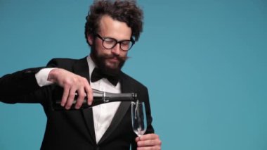 drunk elegant young groom filling empty glass with champagne, measuring the volume by eye and drinking, enjoying, nodding and inviting to drink while smiling on blue background