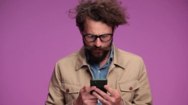 Şok olmuş kıvırcık saçlı adamın cep telefonu tutması, haberleri okuması, şaşırması ve ağız kaplamasının hayal kırıklığına uğraması ve stüdyoda pembe arka planda kafa sallaması.