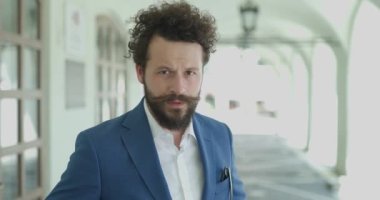 Seksi sakallı adam ceketini ayarlıyor ve düğmeli ceketini ilikliyor, şehrin arka planında kendine güvenen bir şekilde poz veriyor.