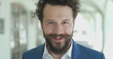 Bıyıklı ve uzun sakallı zeki bir genç adam. Dışarıdaki arka planda gülümsüyor ve mutlu.